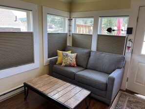 A cozy living room with a grey sofa, two pillows, a wooden coffee table, and a view of the outdoors through large windows with blinds.