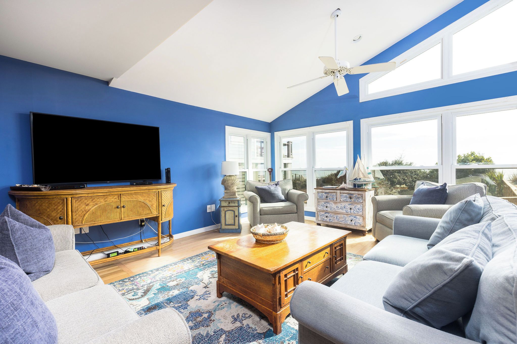 A living room with a flat-screen TV, a ceiling fan, and a large window.