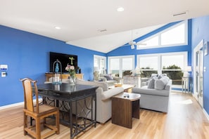 A modern living room with a blue accent wall, a dining area with a wooden table and chairs, and a large window providing natural light. - Stay @ Fun in the Sun! (Folly Beach)