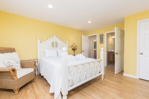 A bedroom with a four-poster bed, a wicker chair, and a bathroom visible through an open door.