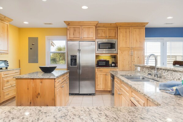 A kitchen with wooden cabinets, stainless steel appliances, granite countertops, and a window with blinds. - Stay @ Fun in the Sun! (Folly Beach)