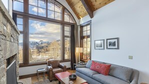 A cozy living room with a stone fireplace, a large window with a mountain view, a grey sofa, and a red bench. - Courcheval D | Luxury Mountain Townhome with Views, Fireplaces & Ski Access (Mountain Village)