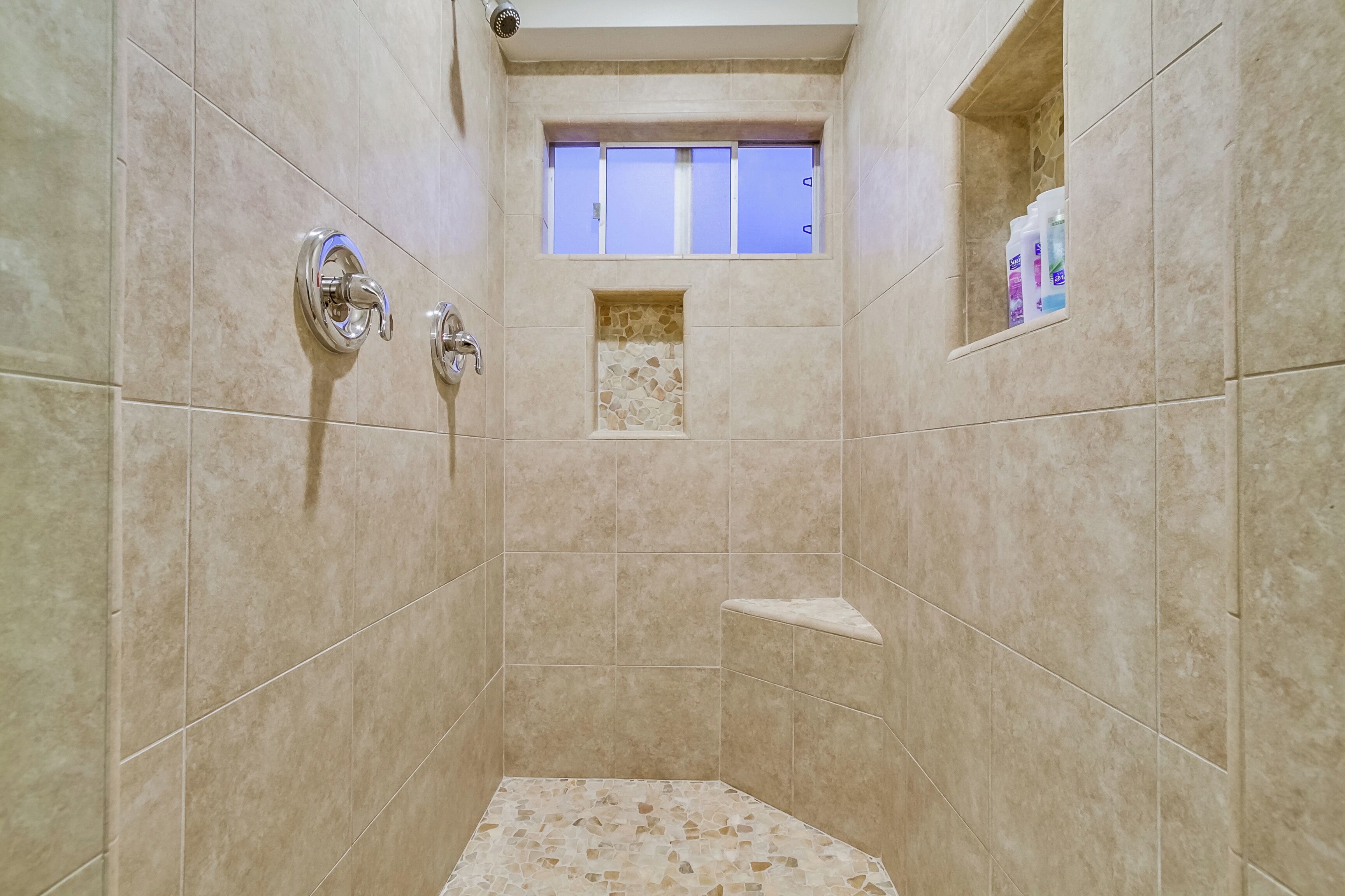Eine Dusche mit beigen Fliesen, einem kleinen Fenster und einem Ablagefach für Pflegeprodukte.