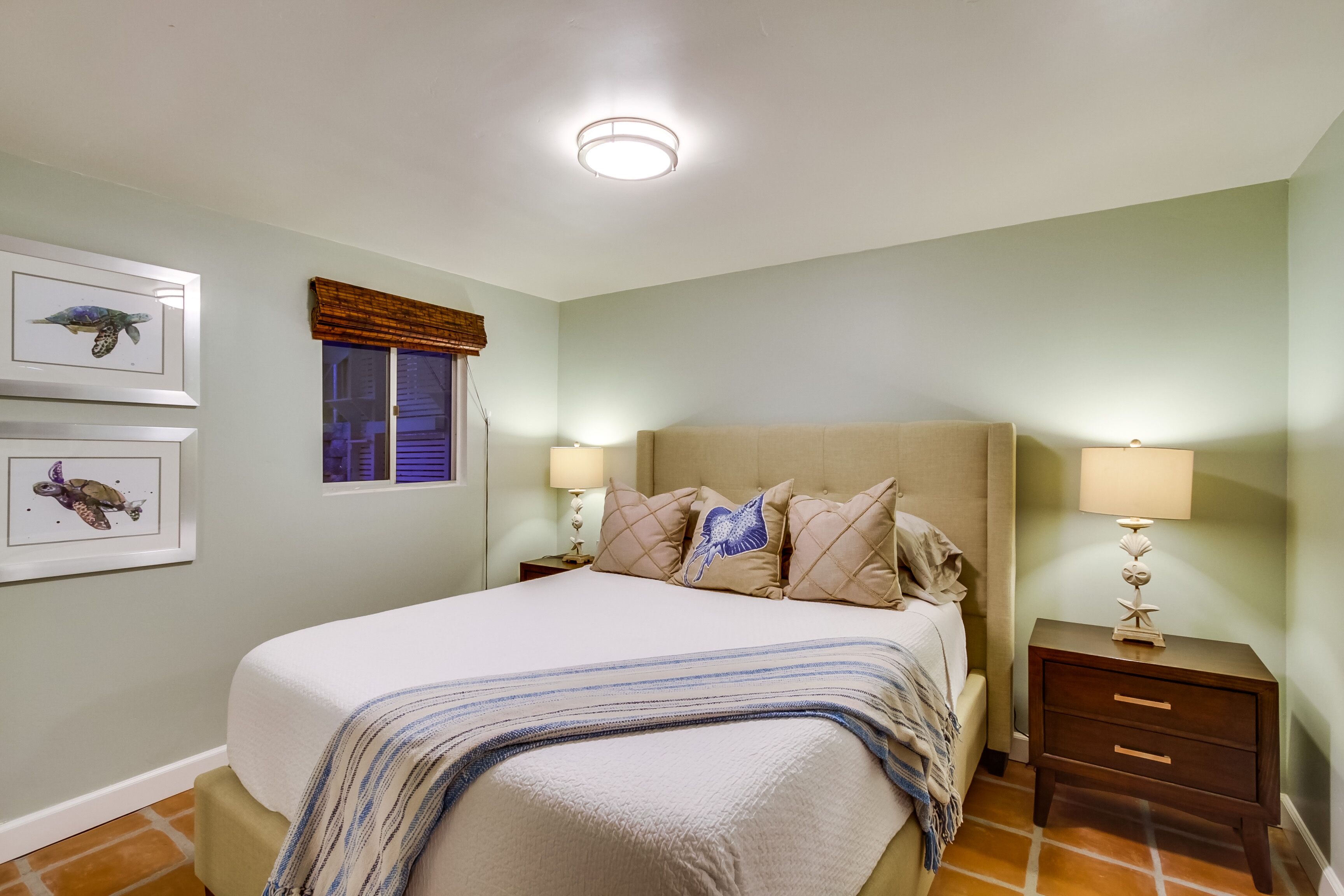 Une chambre à coucher comprenant un lit, deux tables de chevet, une fenêtre avec des stores et deux tableaux encadrés accrochés au mur.