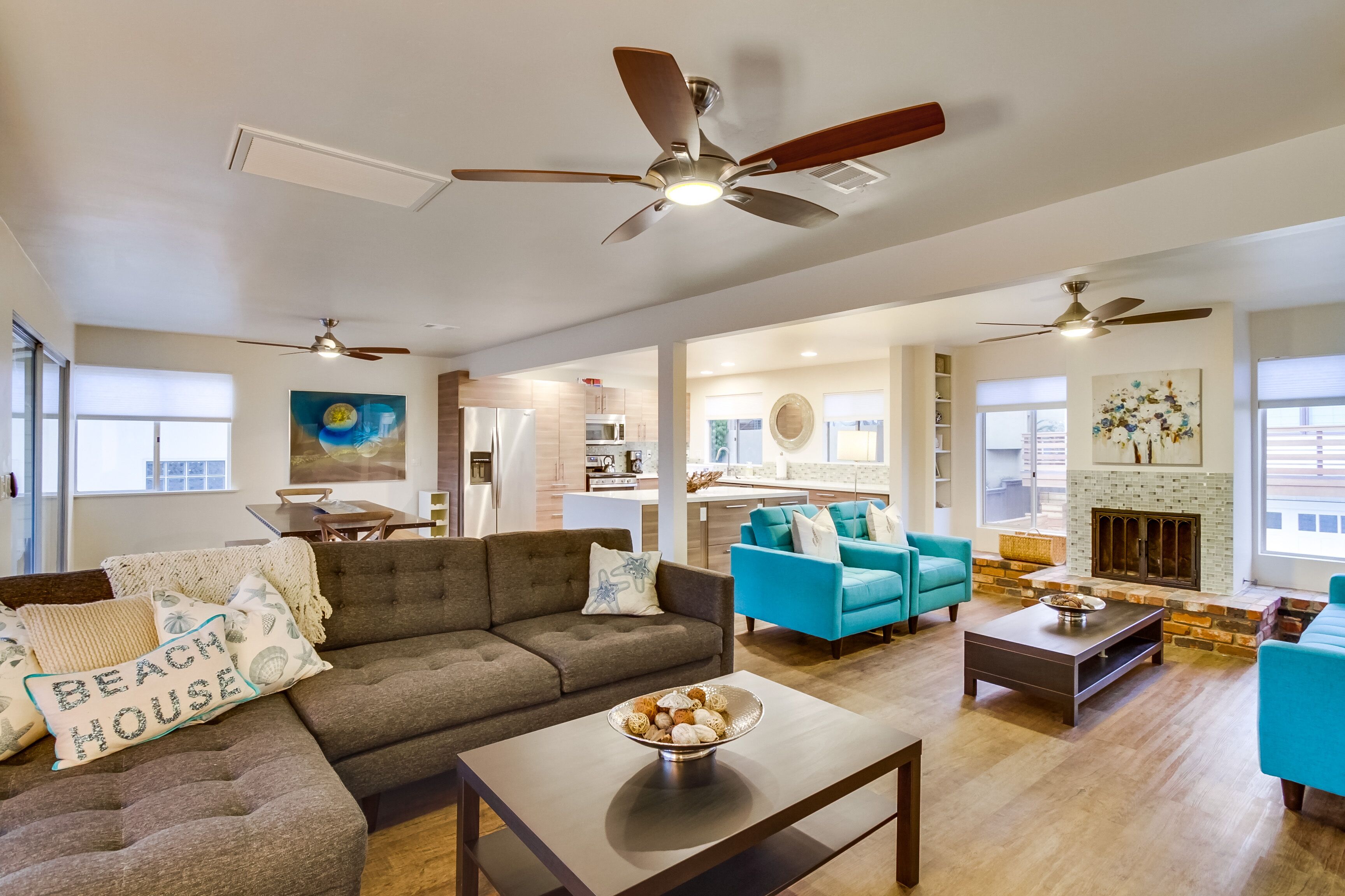 A spacious living room with a grey sofa, blue armchairs, and a coffee table. There are ceiling fans, a fireplace, and a kitchen in the background.