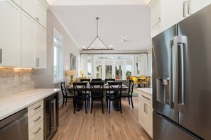 A modern kitchen with white cabinets, a large stainless steel refrigerator, and a dining area with a table and chairs. - Stay @ Pavilion Paradise on Isle of Palms (Isle of Palms)