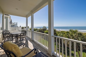 A covered outdoor area with a table and chairs, a view of the beach, and a building with a balcony.