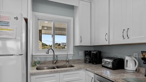 A kitchen with white cabinets, a double sink, a toaster, a coffee maker, and a view of the outdoors.