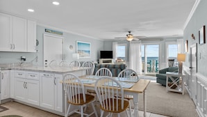 A spacious kitchen with white cabinets, a dining area with white chairs, and a view of the ocean through large windows.