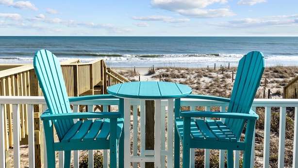 A beachside deck with two turquoise Adirondack chairs and a small table, overlooking the ocean.