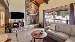 A cozy living room with a stone fireplace, a flat-screen TV, a comfortable sofa, and a balcony with a view of a snowy landscape. - ✨ Newly Remodeled Sleeps 8 Pool, Hot Tubs,Fireplace, Private Deck (Steamboat Springs)