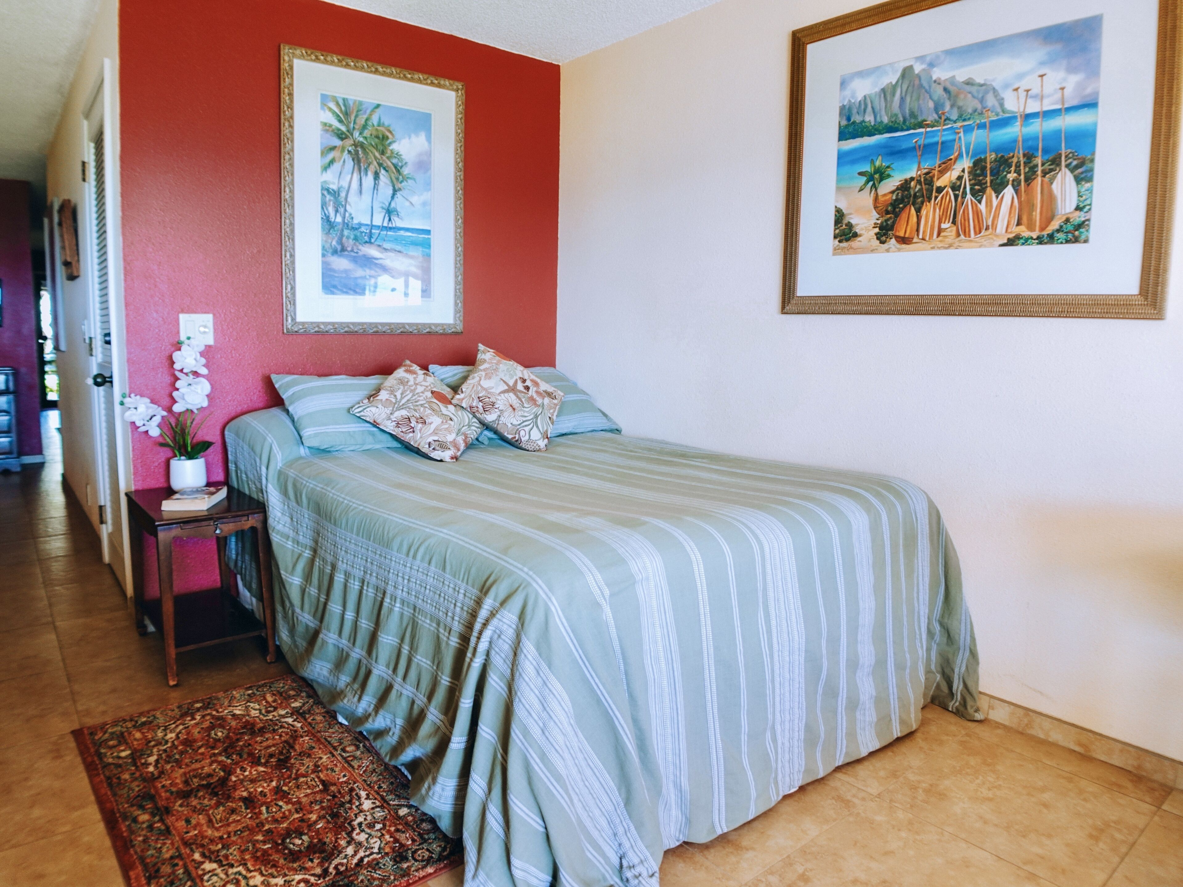 A bedroom with a bed, pillows, a nightstand, a vase of flowers, a rug, and two framed pictures on the wall.