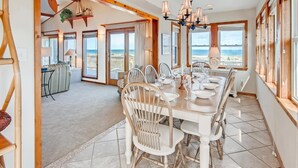 Ein Essbereich mit einem großen Tisch, gedeckt für sechs Personen, umgeben von weißen Stühlen mit Holzbeinen. Der Raum hat große Fenster mit Blick auf den Strand.