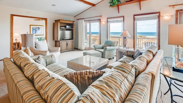 Ein geräumiges Wohnzimmer mit einem großen gestreiften Sofa, einem Fernseher und einem Balkon mit Blick auf den Strand.