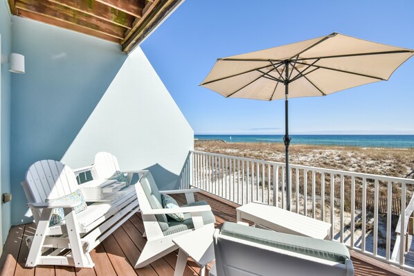 Une terrasse avec des fauteuils Adirondack blancs et une table, un parasol beige, et une vue sur la plage et l’océan.