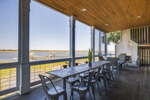 Un vaste espace extérieur aménagé pour les repas, avec vue sur l’eau, comprenant une longue table et de nombreuses chaises, un plafond en bois et de grandes fenêtres.