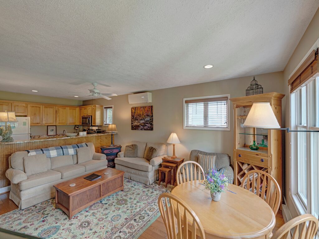A spacious living area with a sofa, coffee table, and dining table. There is a kitchen in the background with wooden cabinets and a window with blinds.