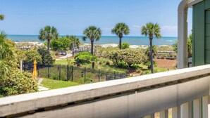 Un balcón con vista a la playa, palmeras y un jardín cercado.