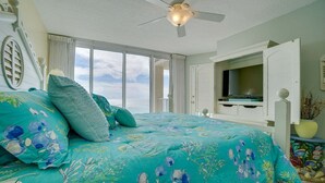 A bedroom with a large bed, a ceiling fan, a TV, and a view of the ocean.