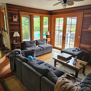 A living room with a sofa, a coffee table, and a television. There is a window with a view of a pool and greenery outside.