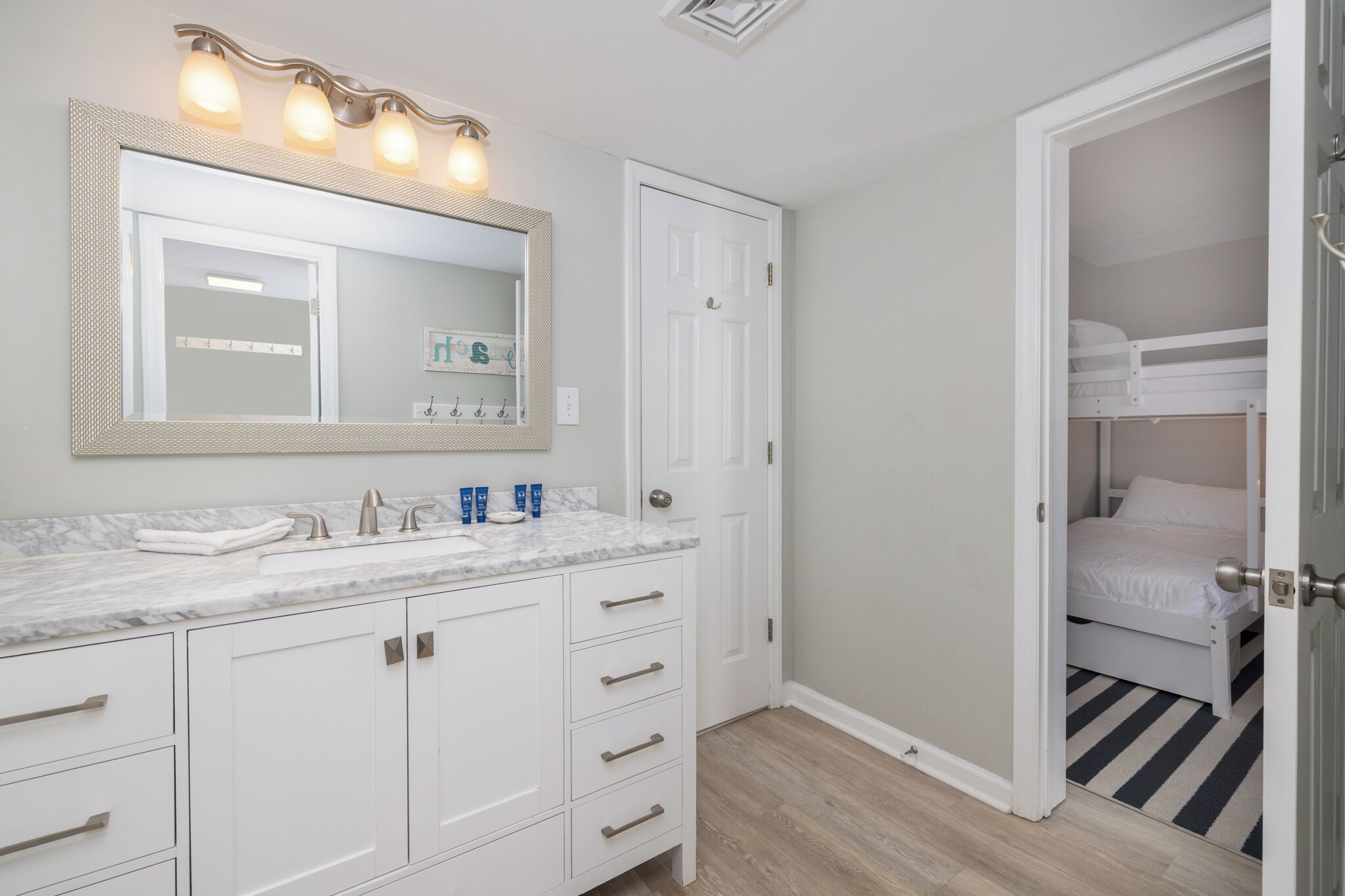 A bathroom with a large mirror, a sink with toiletries, and a door leading to a bedroom with a bunk bed.