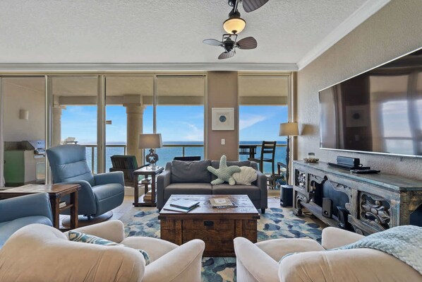 A spacious living room with a large TV, comfortable seating, a ceiling fan, and a view of the ocean.