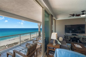 A balcony with a view of the beach and ocean, a seating area with a TV and speakers, and a ceiling fan.