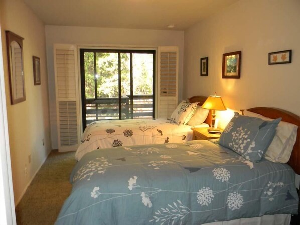 Un dormitorio con dos camas, una ventana con contraventanas, una lámpara de noche y cuadros en la pared.