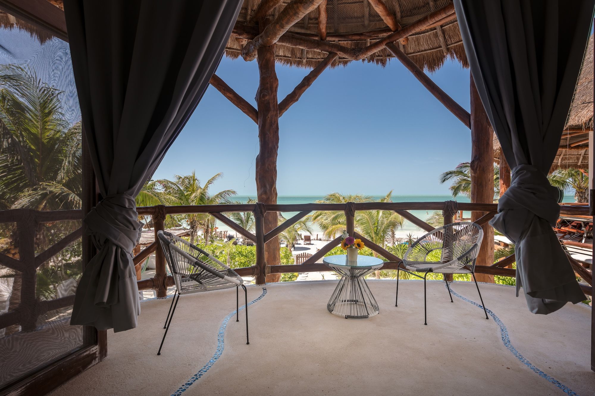 Une vue sur la plage depuis une terrasse en bois dotée d’un toit de chaume, d’un hamac et d’une table ornée d’un vase de fleurs.