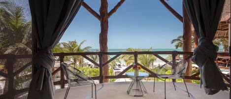A beach view from a wooden deck with a thatched roof, a hammock, and a table with a vase of flowers.