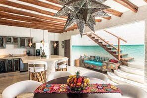 A modern kitchen with a central island, a dining area with a fruit bowl, and a view of the sea through a glass wall.