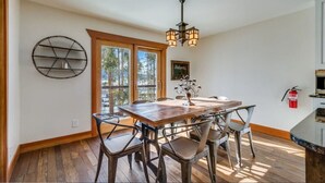 Un coin repas avec une table en bois et des chaises en métal, une étagère murale ronde, un lustre et une fenêtre donnant sur des arbres.