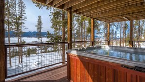 Une terrasse en bois avec un jacuzzi, une rambarde en métal et une vue sur un paysage enneigé, avec des arbres et du sol recouverts de neige.