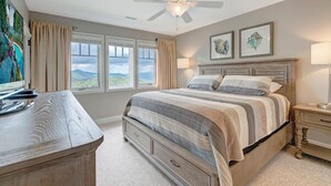 Une chambre avec un grand lit, un ventilateur de plafond, deux fenêtres avec des rideaux, un bureau et des œuvres d’art accrochées au mur.