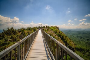 A long, narrow wooden bridge with metal railings, leading to a viewpoint overlooking a mountainous landscape with dense green forests and a clear blue sky. - Townhome R5B- Mtn Home/View, Foosball, Indoor/Outdoor Pools & Hot Tub, Fireplace (Banner Elk)