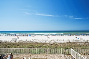 Una spiaggia con sabbia bianca, cielo azzurro limpido e mare. Si vedono ombrelloni e persone sulla spiaggia.
