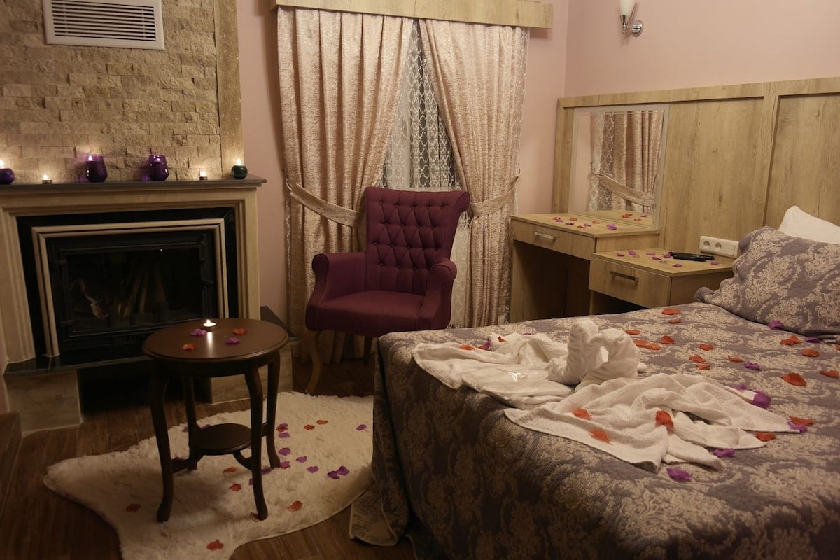 A cozy bedroom with a fireplace, a tufted armchair, a bed with rose petals, and a wooden desk.