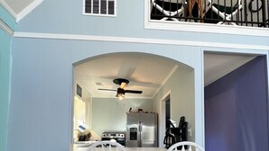Una cocina con una isla central, dos taburetes blancos tipo barra, un ventilador de techo y vistas del nivel superior a través de un ventanal.