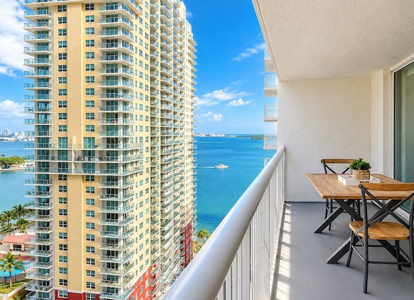 A high-rise building with a balcony featuring a table and chairs, overlooking a body of water with a distant cityscape.
