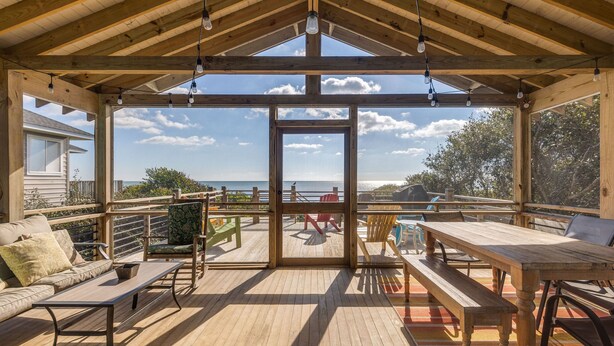 Eine Holzterrasse mit Holztisch und -stühlen, einer Sitzecke und Blick auf den Ozean.