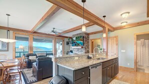 A modern kitchen with granite countertops, dark cabinetry, and a large island. There is a dining area with a table and chairs, and a view of the mountains through large windows. - Chalakee Lodge CH-3- Mountain View, Gated Resort, Indoor/Outdoor Pools & Hot Tub (Boone)
