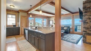 A spacious kitchen with dark cabinets, a granite countertop, and pendant lighting. A large island with a sink and a dining area with a view is visible. - Chalakee Lodge CH-3- Mountain View, Gated Resort, Indoor/Outdoor Pools & Hot Tub (Boone)
