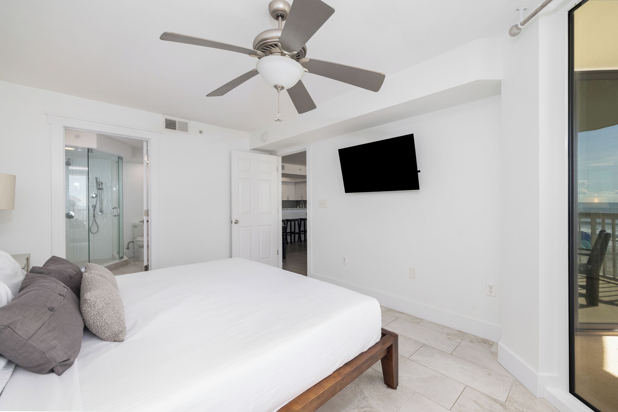 Une chambre à coucher avec un lit, un ventilateur de plafond, un téléviseur fixé au mur et une vue sur le balcon.