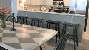 Eine moderne Küche mit weißen Schränken, einer Mikrowelle, einem Herd und einem Kühlschrank. Ein karierter Tisch steht mit Stühlen und einer Blumenvase bereit.