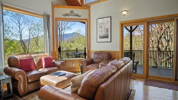 Ein geräumiges Wohnzimmer mit Ledersofas, einem Couchtisch und großen Fenstern mit Blick auf Bäume und Berge.