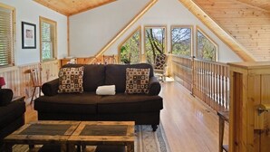 Ein gemütliches Loft-Zimmer mit Holzboden, einem dunkelbraunen Sofa mit gemusterten Kissen, einem Holztisch und Blick auf Bäume durch große Fenster.