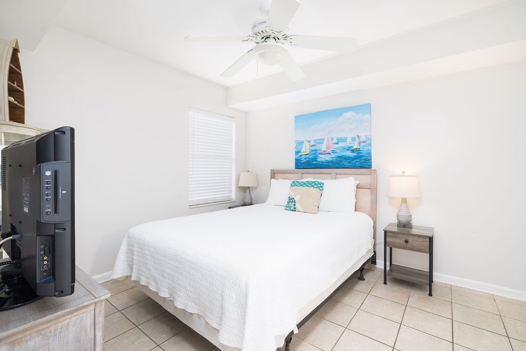 Une chambre à coucher comprenant un lit, un ventilateur de plafond, un tableau représentant des voiliers, un téléviseur et une table de chevet avec une lampe.
