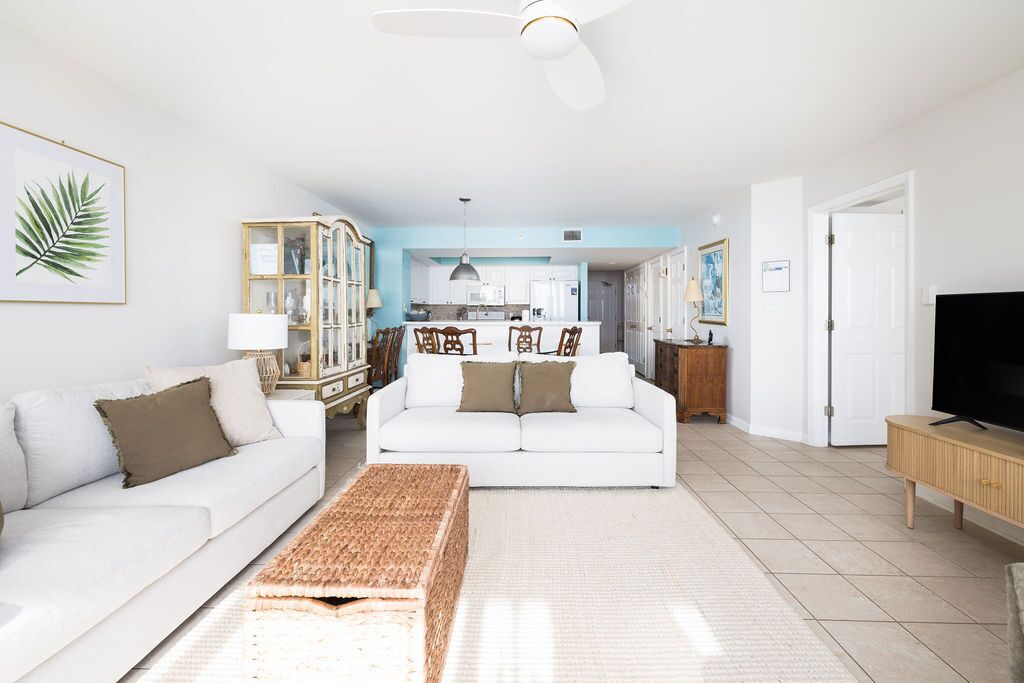 A living room with a white sofa, a coffee table, and a television. There is a kitchen area in the background with white cabinets and a blue accent wall.