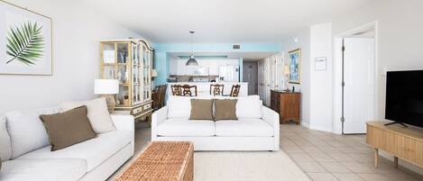 A living room with a white sofa, a coffee table, and a television. There is a kitchen area in the background with white cabinets and a blue accent wall.