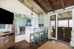 A kitchen with a large island, a dining area with wicker chairs, and a sliding glass door leading to a balcony with a view of the sea.